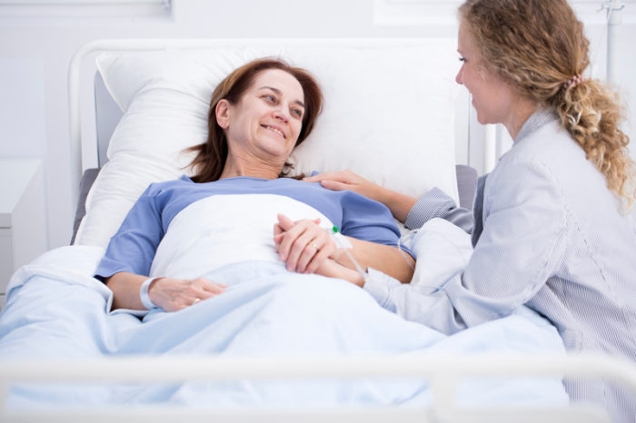 Kvinnlig strokepatient ligger i sjukhussäng och håller sin dotter, som sitter bredvid, i handen.